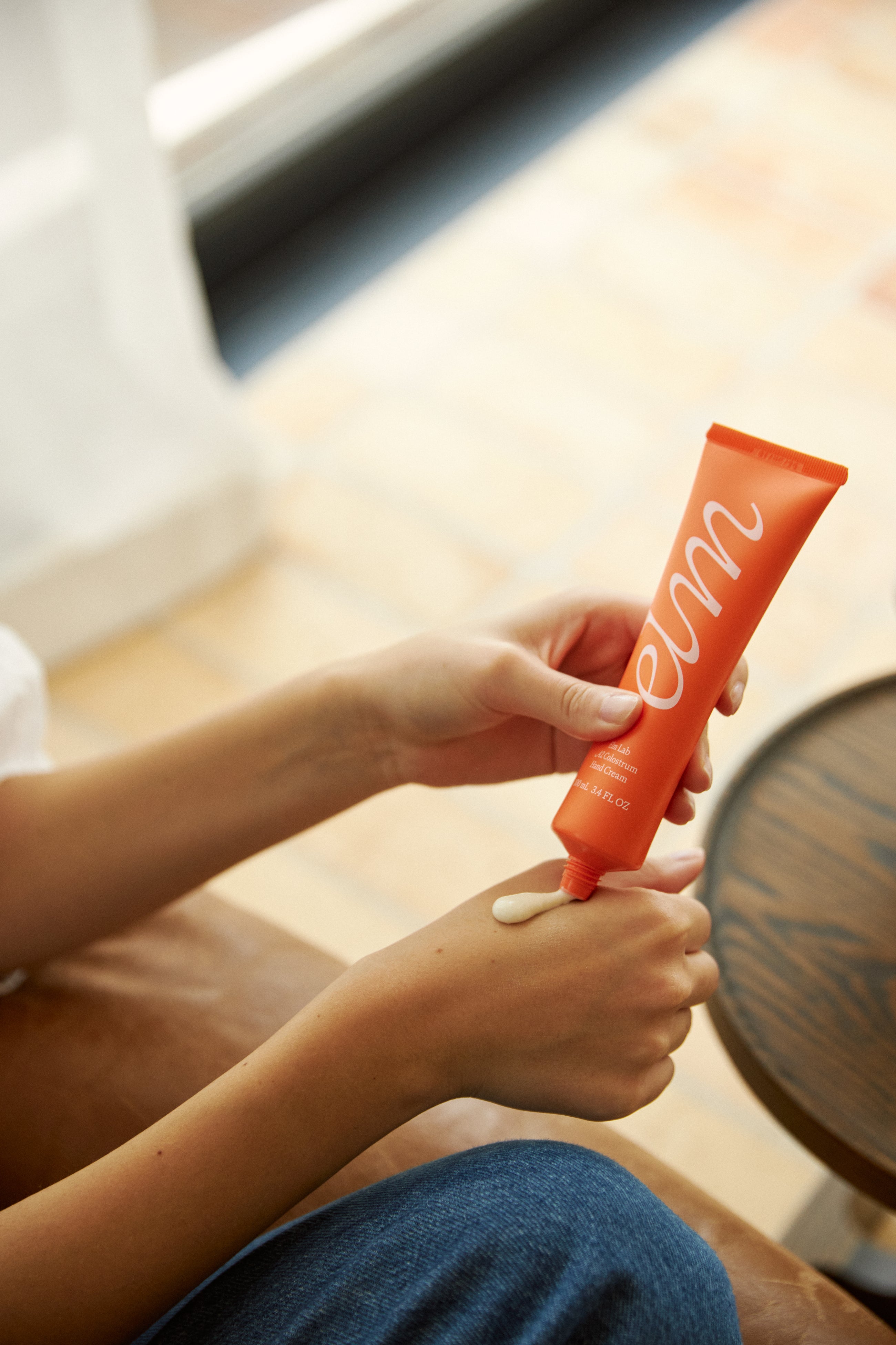 Person applying orange tube of elm hand cream to their arm with a blurred background