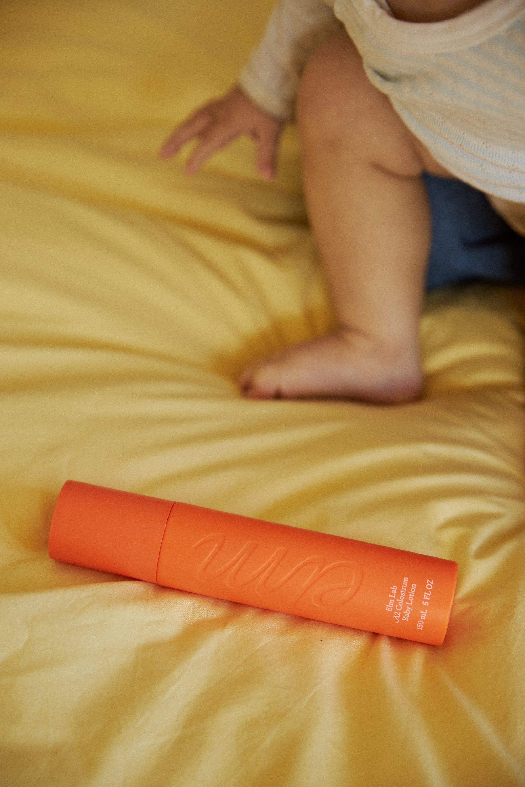 Orange baby lotion on a bed with a child's legs in the background